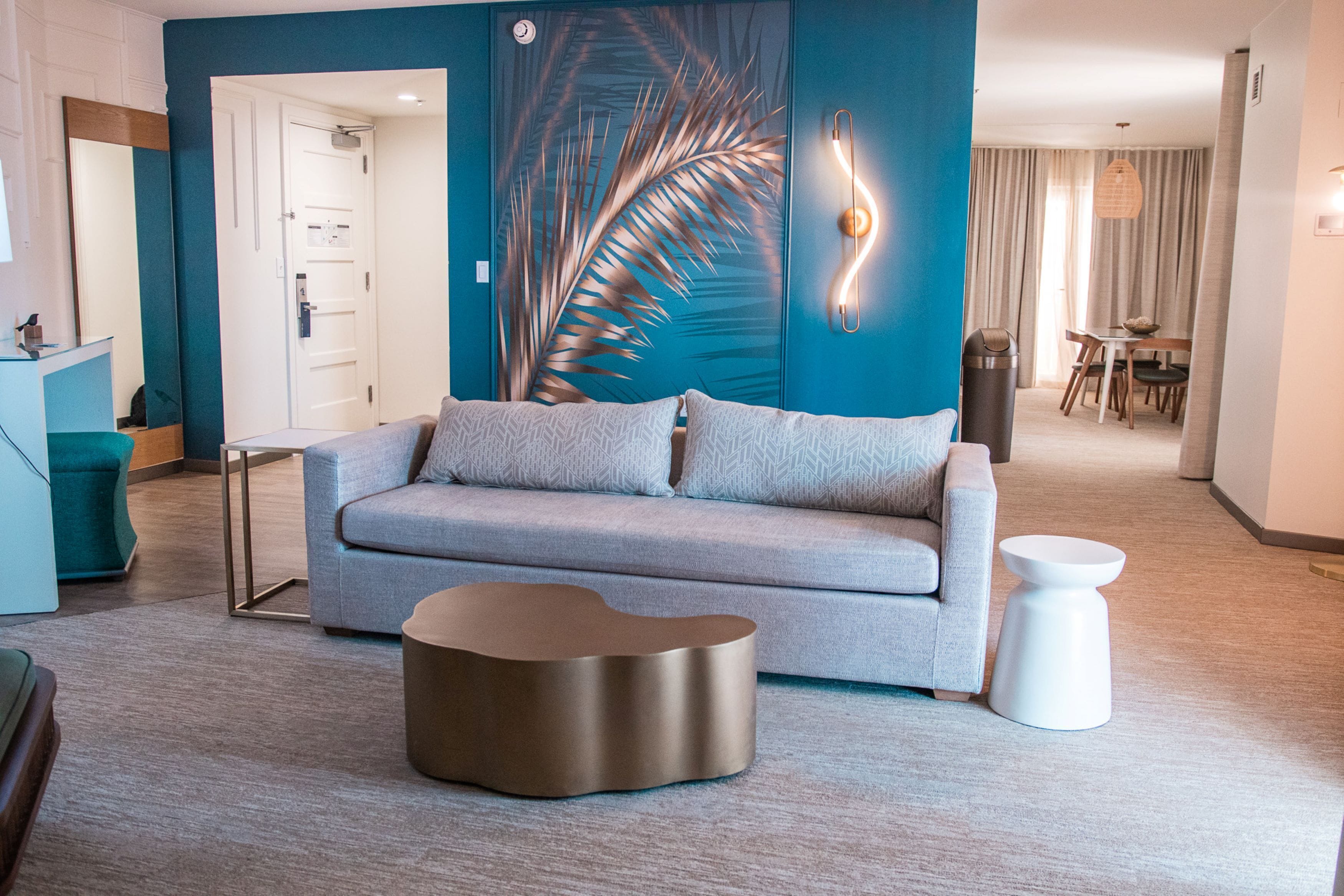 A modern living room with a grey sofa, a unique coffee table, and a wall with a palm leaf design.