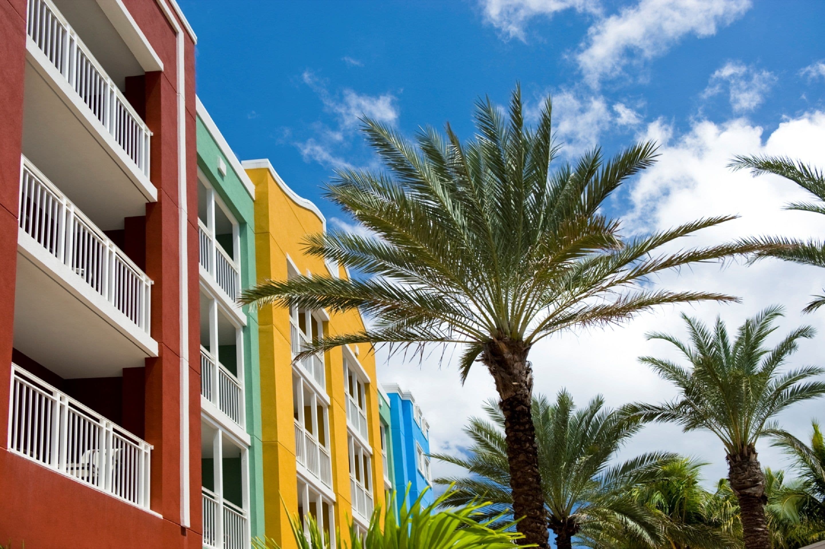 A row of colorful multi-story buildings with balconies, palm trees, and a clear blue sky.