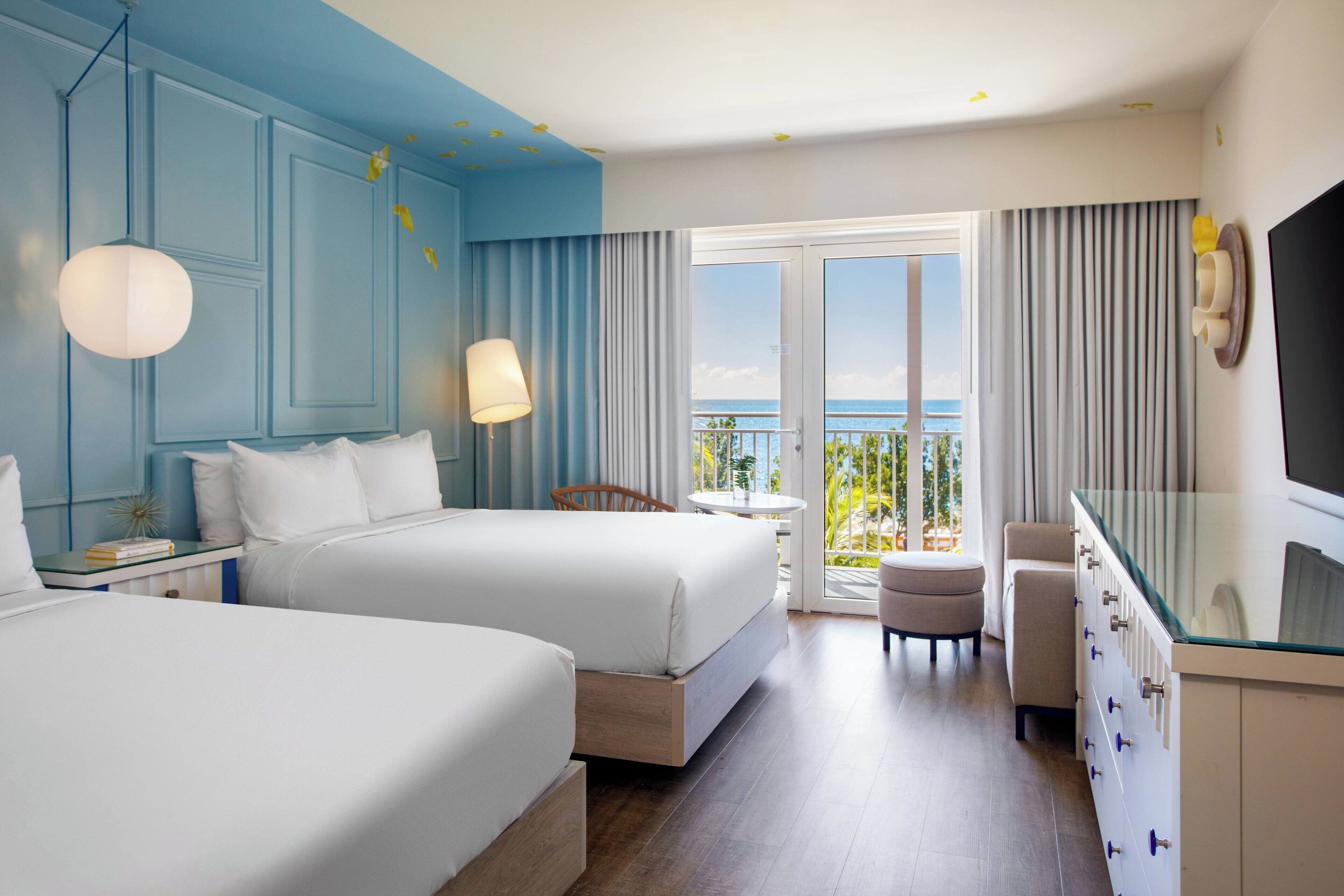 A hotel room with two beds, a TV, a balcony with curtains, and a view of the sea.