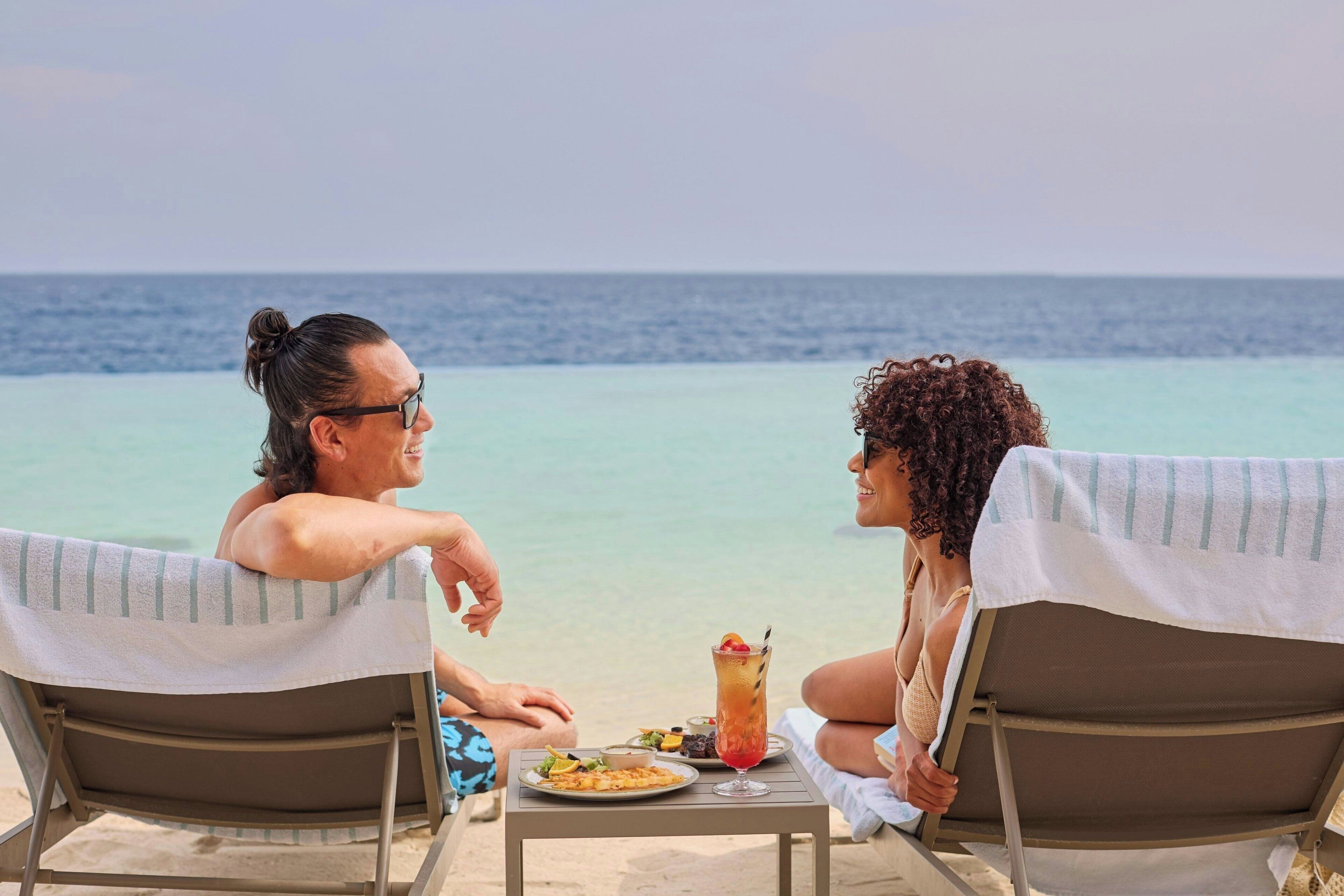 Two people seated at a table with a drink and a plate of food, overlooking a clear blue sea.
