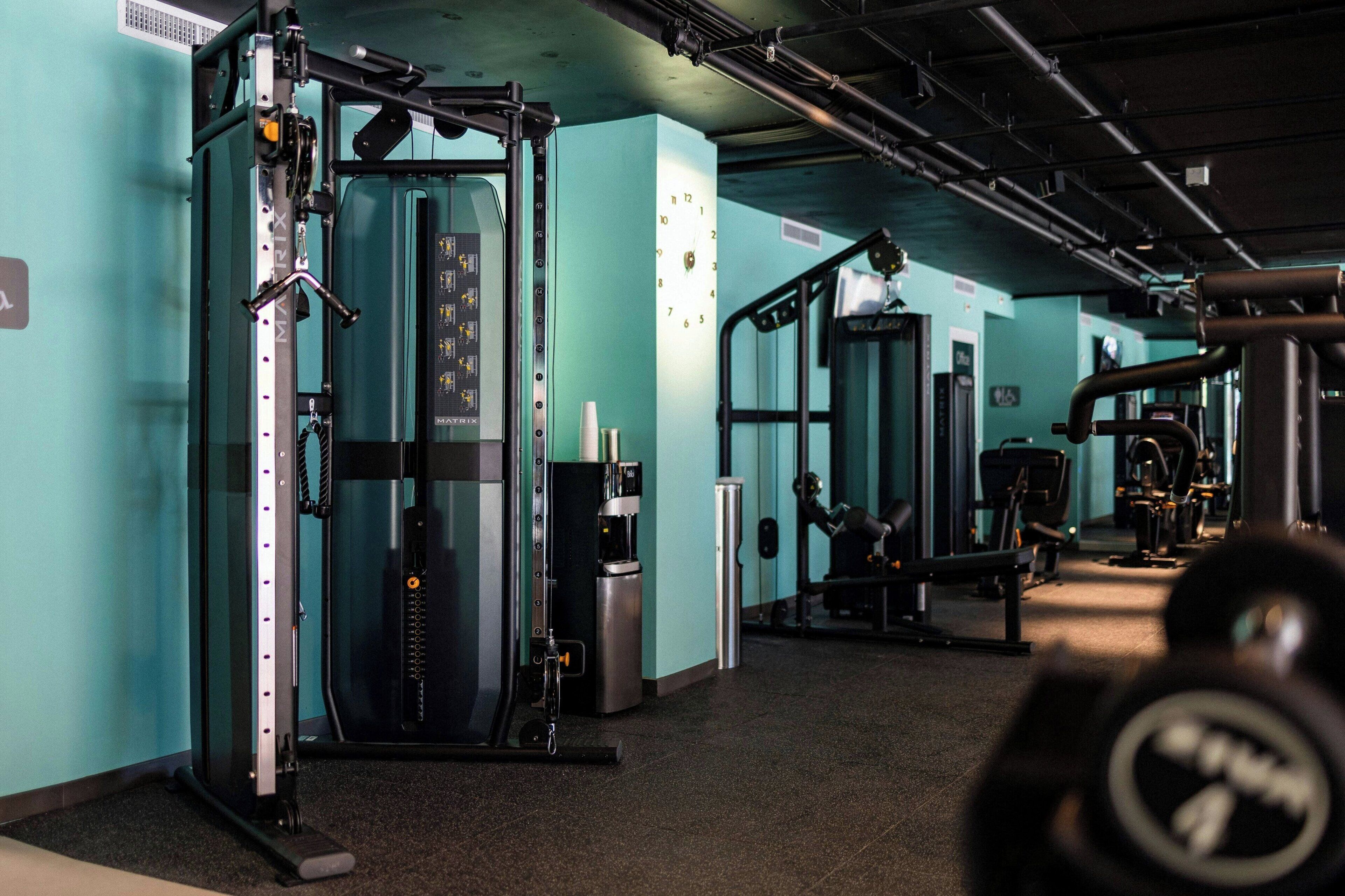 Un gimnasio con diferentes máquinas de ejercicio, un dispensador de agua y un reloj en la pared.