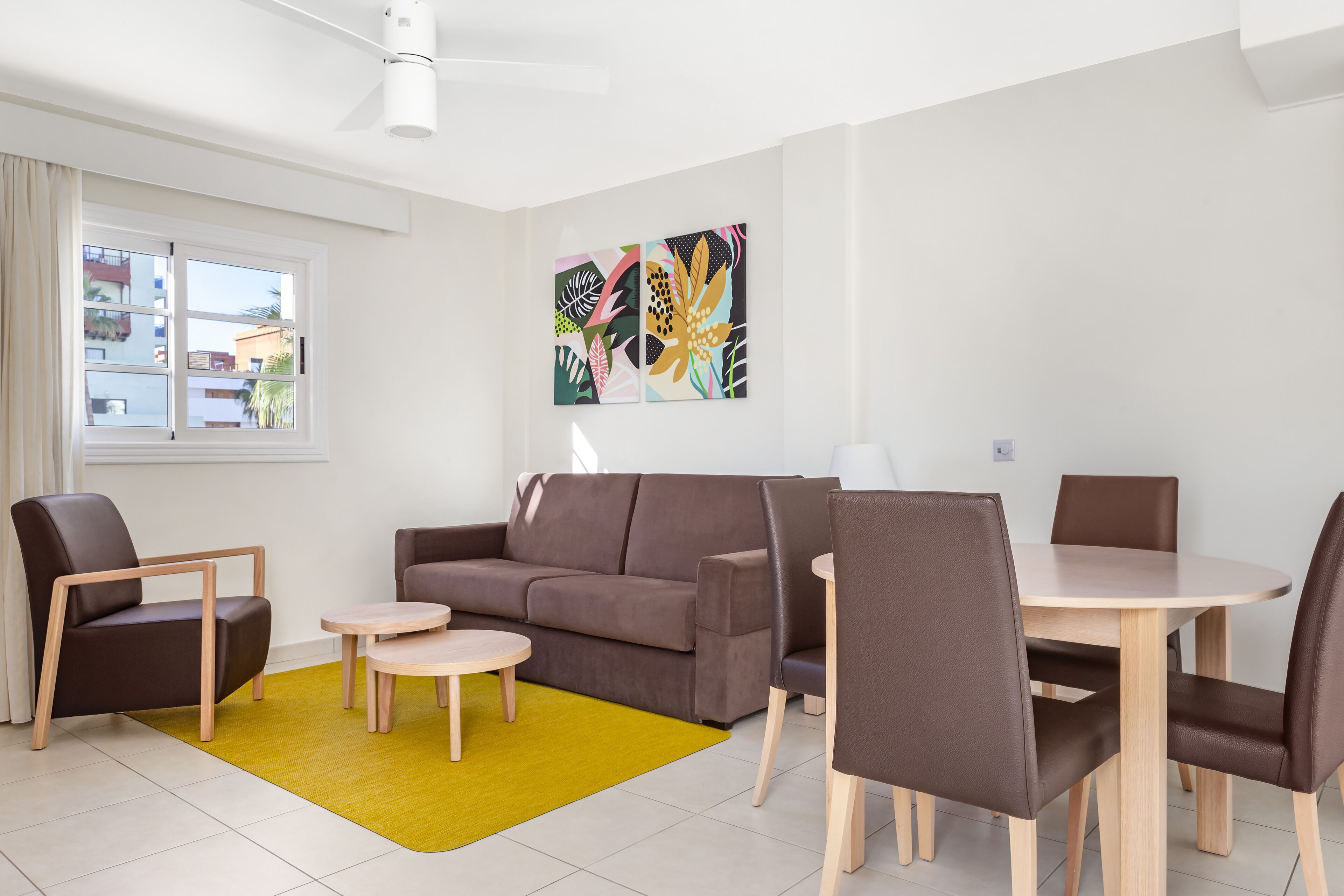 A modern living room with a brown sofa, two armchairs, a small round table, and a dining table with chairs. There is a yellow rug on the floor, and a window with curtains is visible.