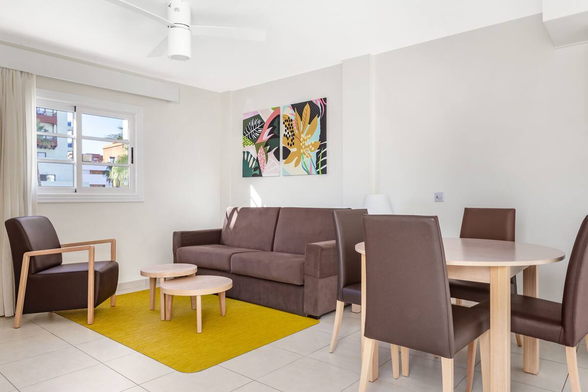 A modern living room with a brown sofa, two armchairs, a small round table, and a dining table with chairs. There is a yellow rug on the floor, and a window with curtains is visible.