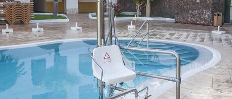 A swimming pool with a safety chair, metal railings, and a tiled deck.