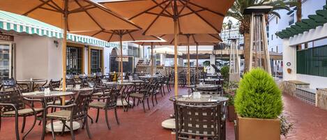 An outdoor café with multiple tables and chairs, umbrellas, and a potted plant.