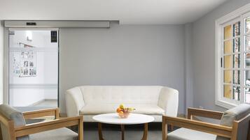Una sala de estar moderna con un sofá blanco, dos sillas de madera, una mesa redonda de centro con un frutero y una ventana con persianas.