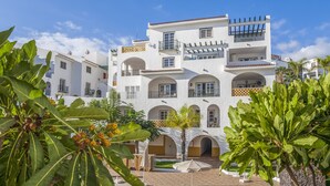 A white multi-story building with balconies, arched doorways, and a courtyard with plants and umbrellas.