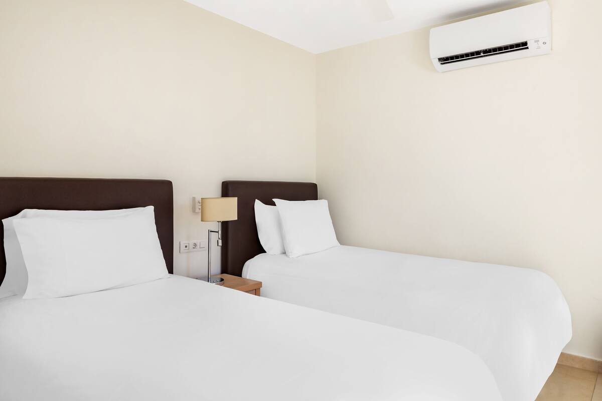 A hotel room with two single beds, a nightstand with a lamp, and a wall-mounted air conditioner.