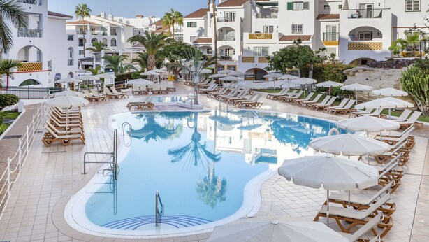 A pool area with lounge chairs and umbrellas, surrounded by white buildings with balconies and palm trees.