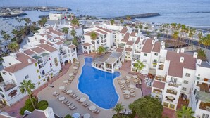 A resort with a central swimming pool surrounded by sun loungers and palm trees, overlooking a marina and the sea.