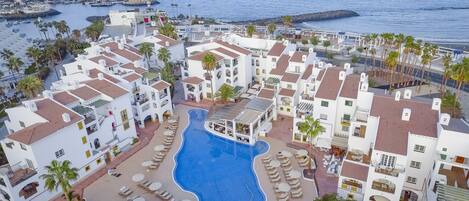 A resort with a central swimming pool surrounded by sun loungers and palm trees, overlooking a marina and the sea.