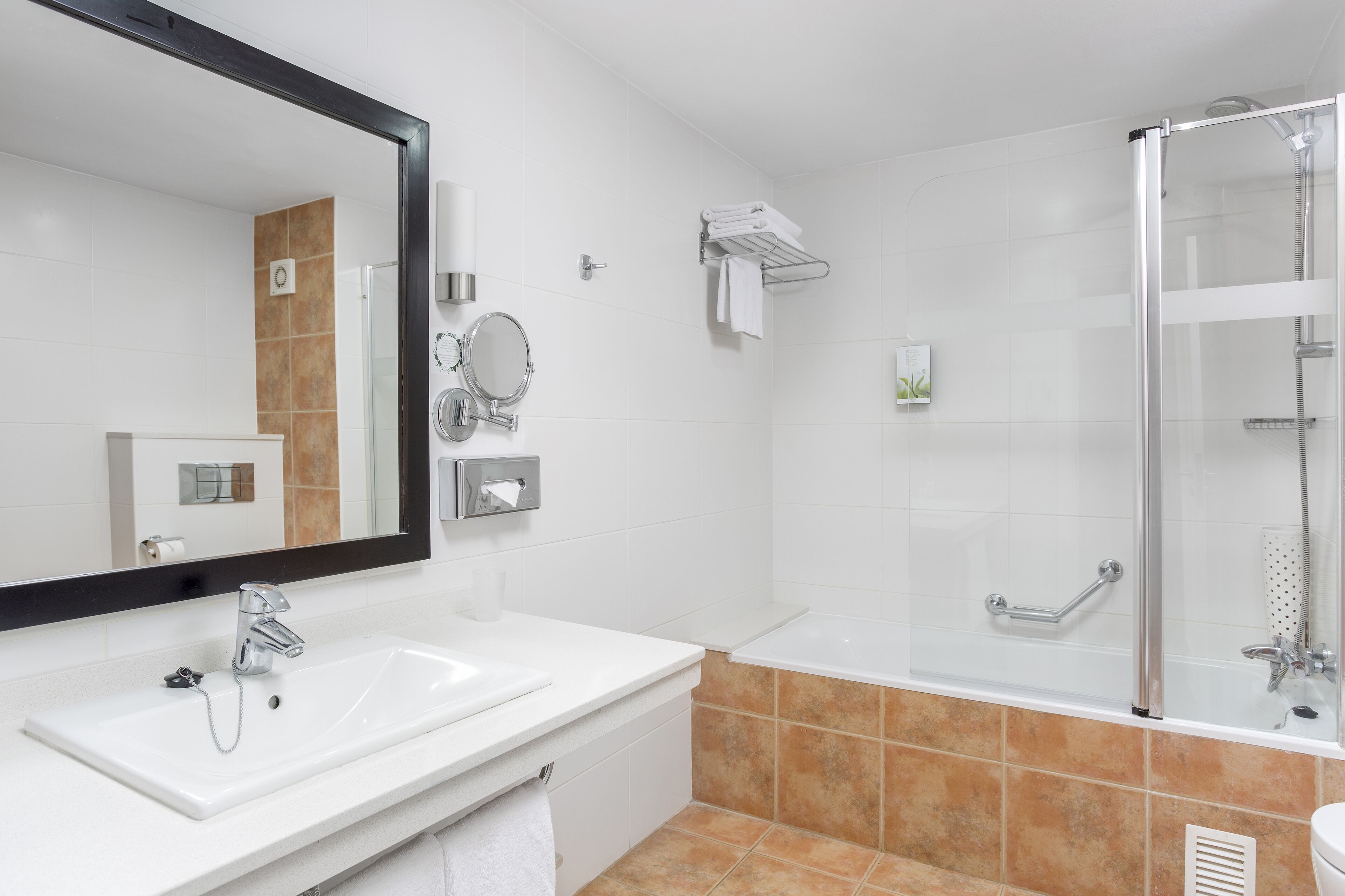 Un baño moderno con lavamanos blanco, un espejo grande, ducha con mampara de vidrio y toallero.