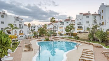 Una piscina rodeada de sillones de descanso y palmeras, con edificios blancos al fondo.