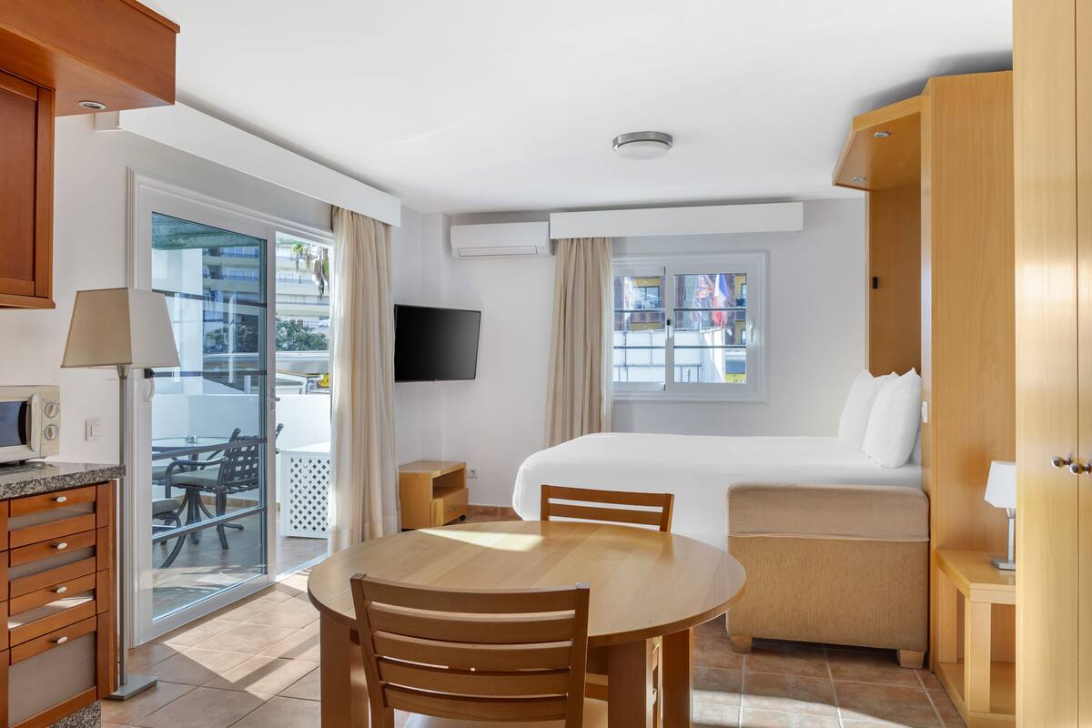 A hotel room with a bed, a dining table with chairs, a TV, and a sliding glass door to the balcony.