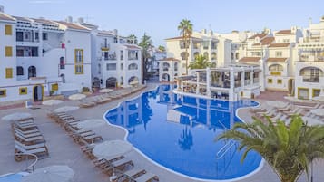 A pool area with sun loungers and umbrellas, surrounded by white buildings with balconies.