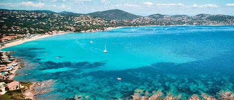An aerial view of a coastal town with clear turquoise waters, a sandy beach, and a hilly backdrop.