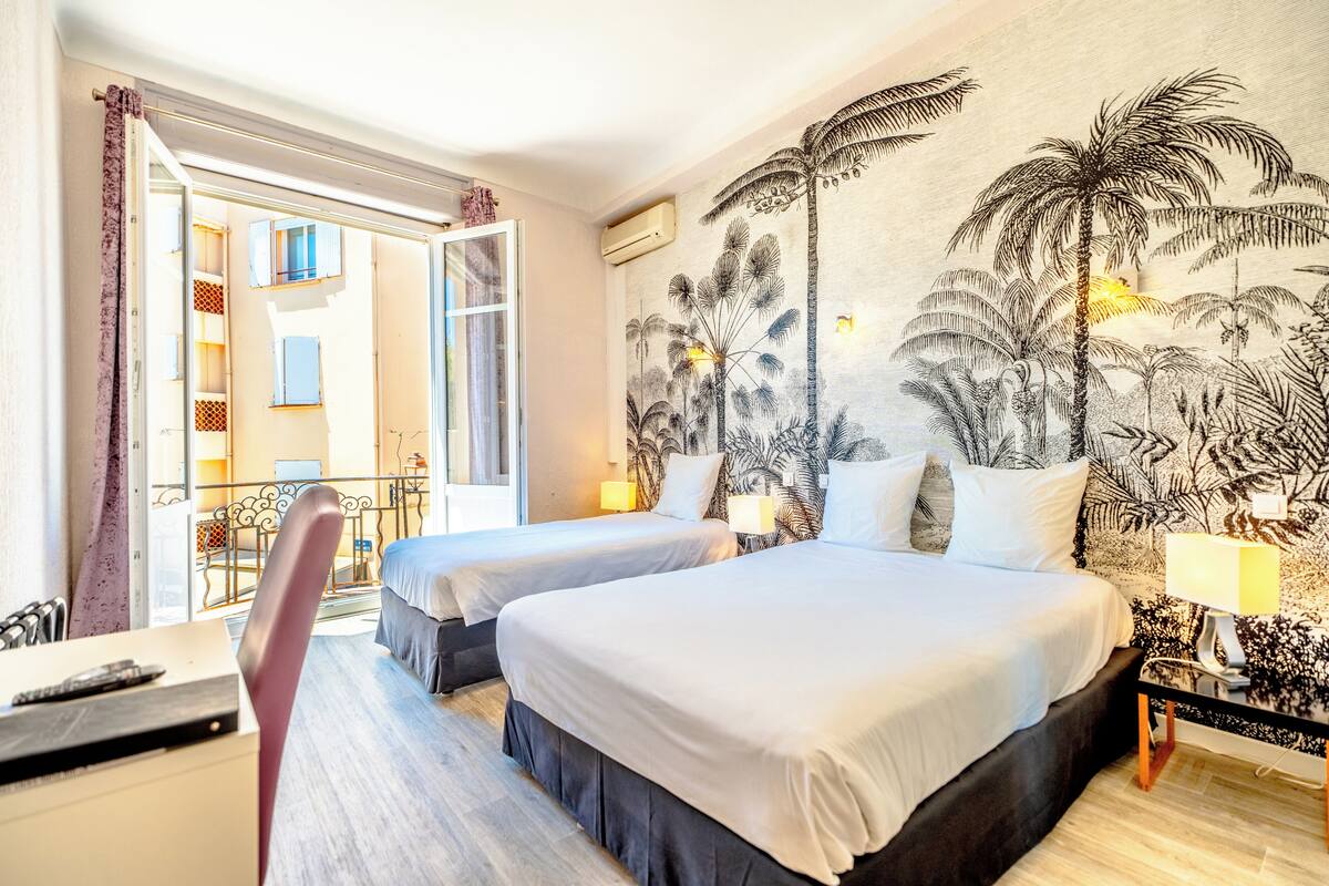 A hotel room with two beds, a desk, and a chair. There is a balcony with a view of buildings.