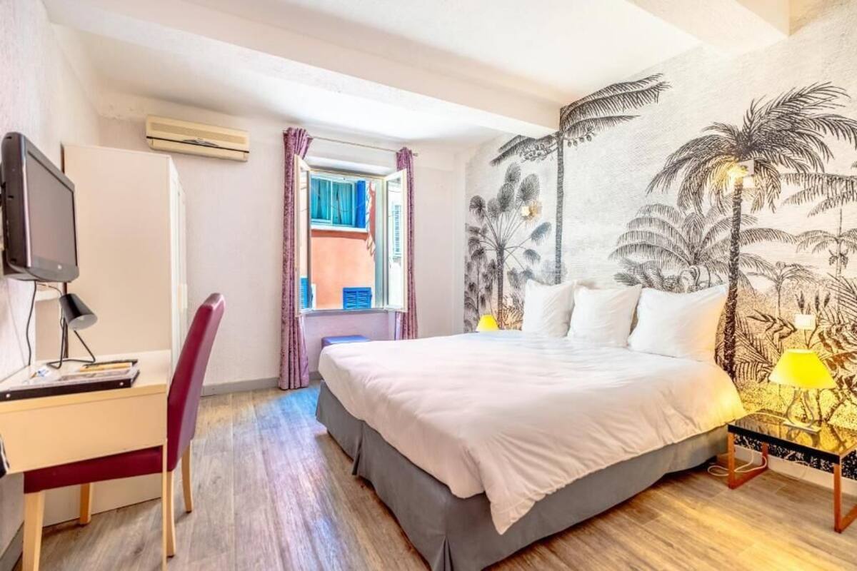 A bedroom with a bed, a desk with a chair, a TV, and a wall with a tropical mural.