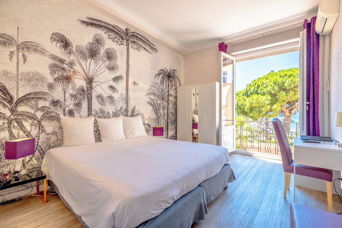 A bedroom with a large bed, a desk, and a view of a balcony with trees.