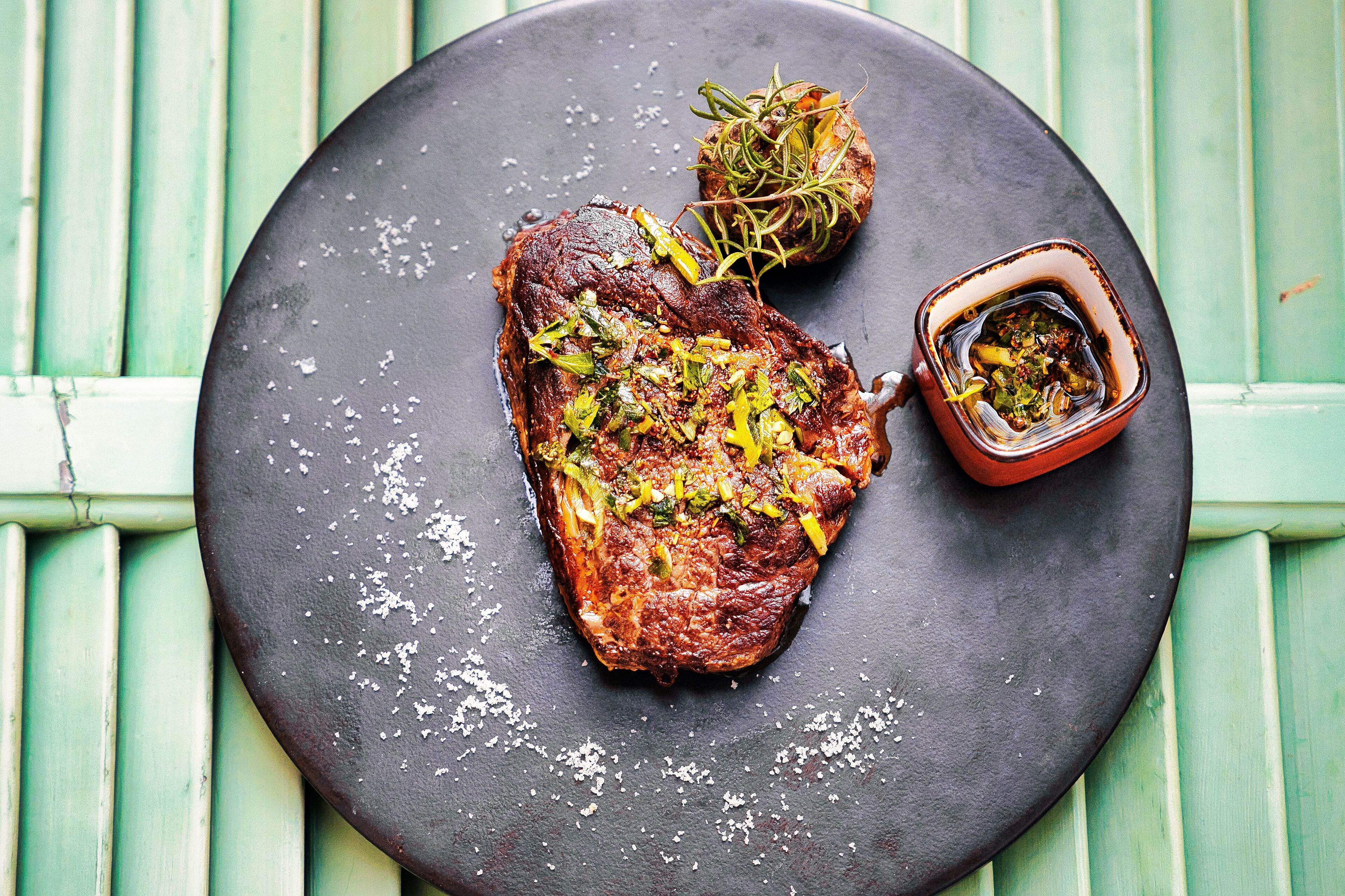 Een steak met garnering op een zwart bord, vergezeld van een kleine schaal saus, voor een groene houten achtergrond.