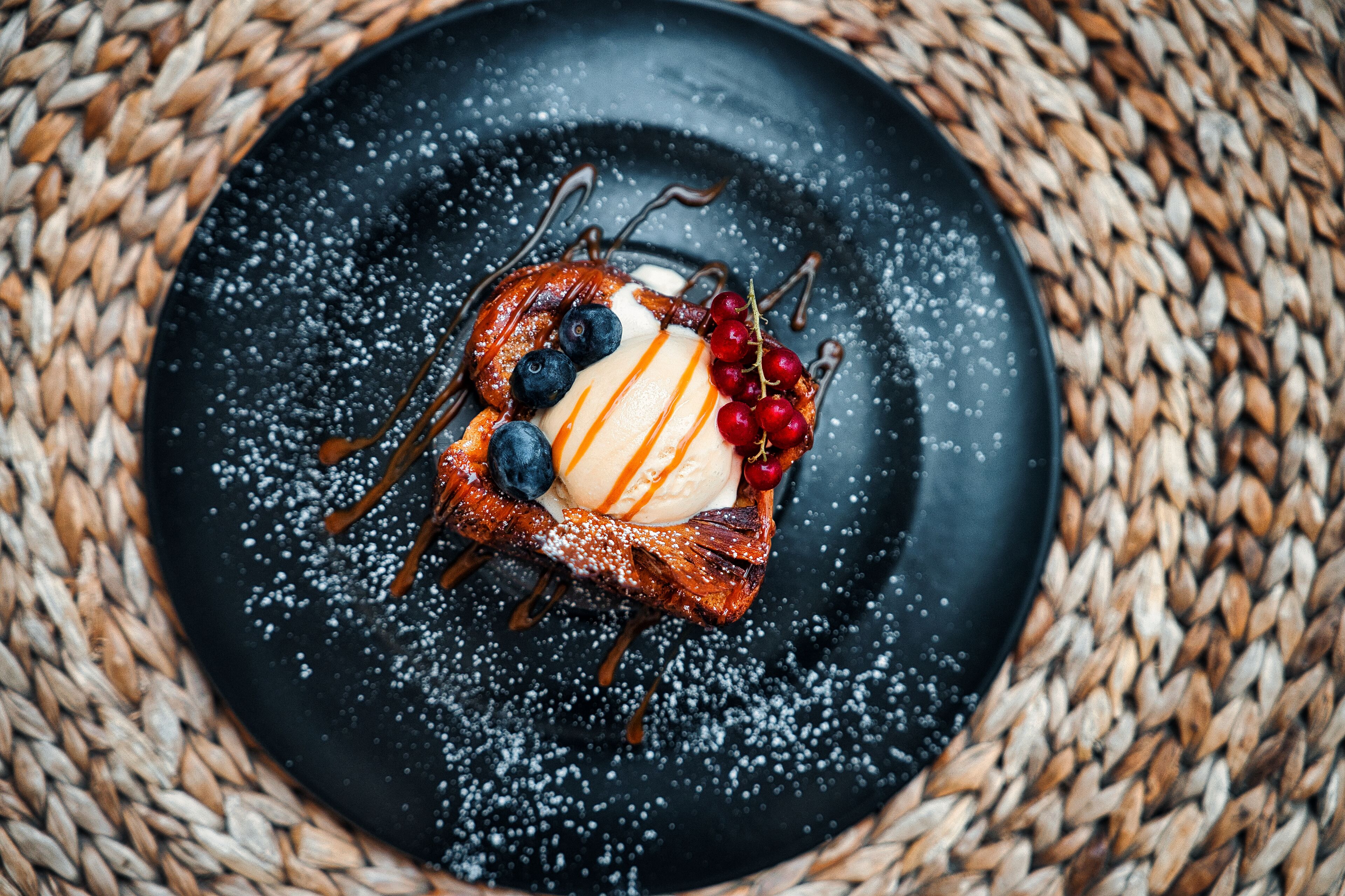 Postre sobre un plato negro con arándanos, cerezas y un hilo de caramelo, colocado sobre un tapete tejido.
