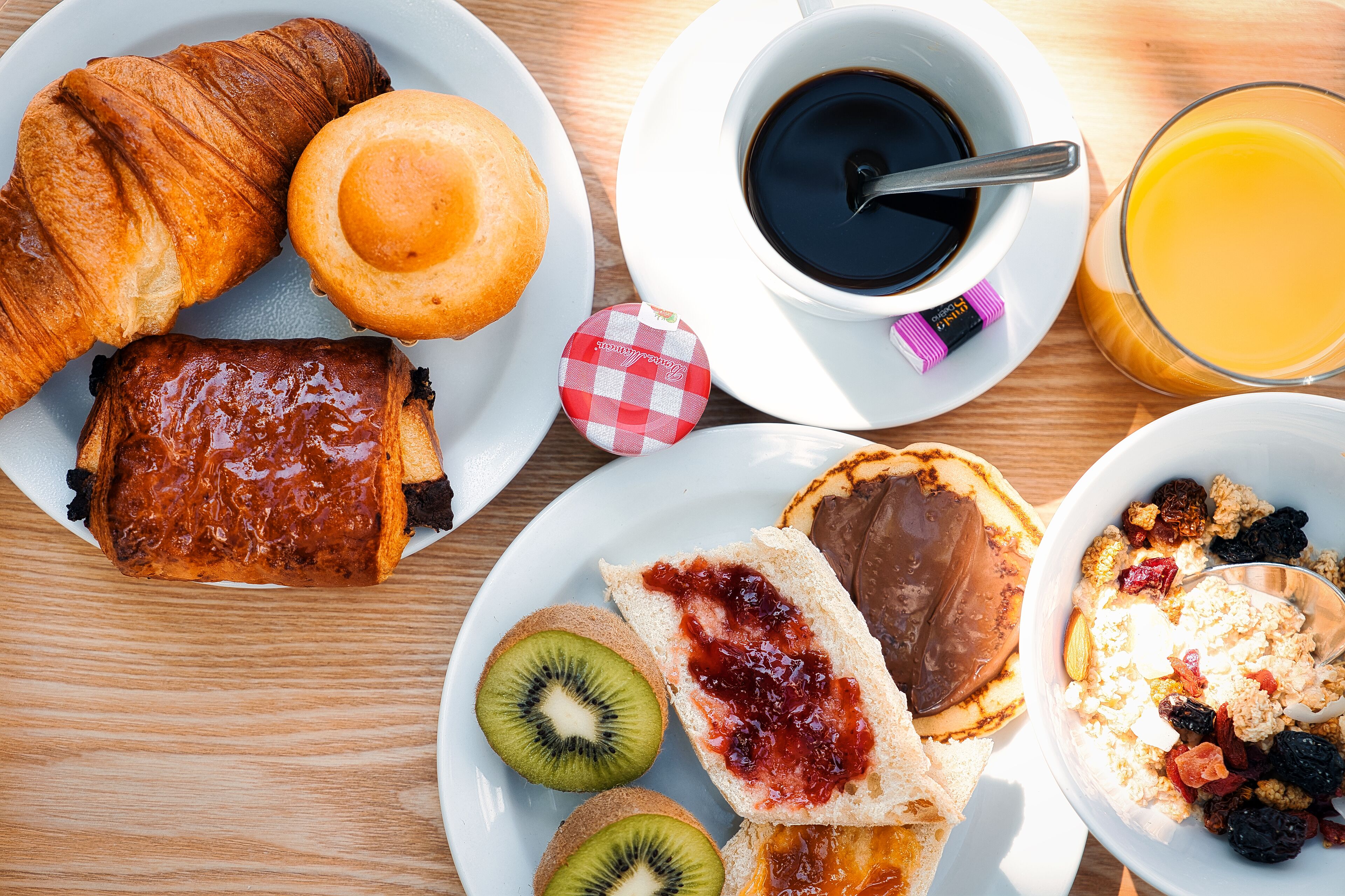A breakfast spread with croissants, a muffin, a chocolate-covered pastry, toast with jam and chocolate, kiwi slices, and a bowl of cereal with nuts and dried fruits.