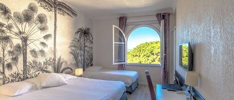 Une chambre d’hôtel avec deux lits, un bureau avec une lampe, une télévision et une fenêtre donnant sur un espace verdoyant.
