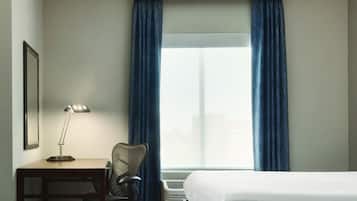 A hotel room with a bed, a desk with a lamp, a chair, and a window with curtains.
