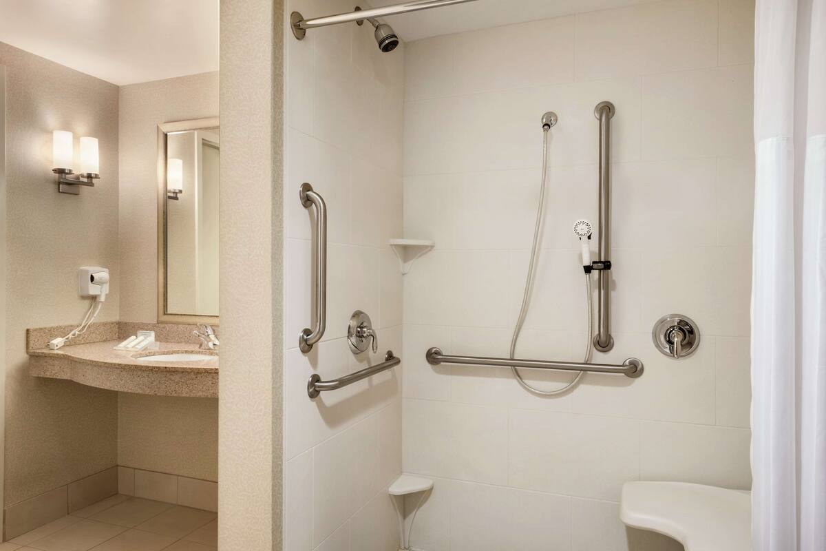 Un baño con lavamanos, espejo y zona de ducha equipada con barras de apoyo y un cabezal de ducha manual.