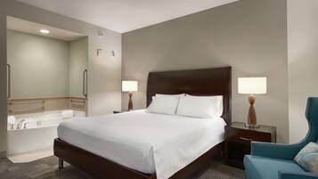 Habitación de hotel con una cama grande, un área de descanso con una silla, una mesita auxiliar, una lámpara, un baño con bañera y un espejo.