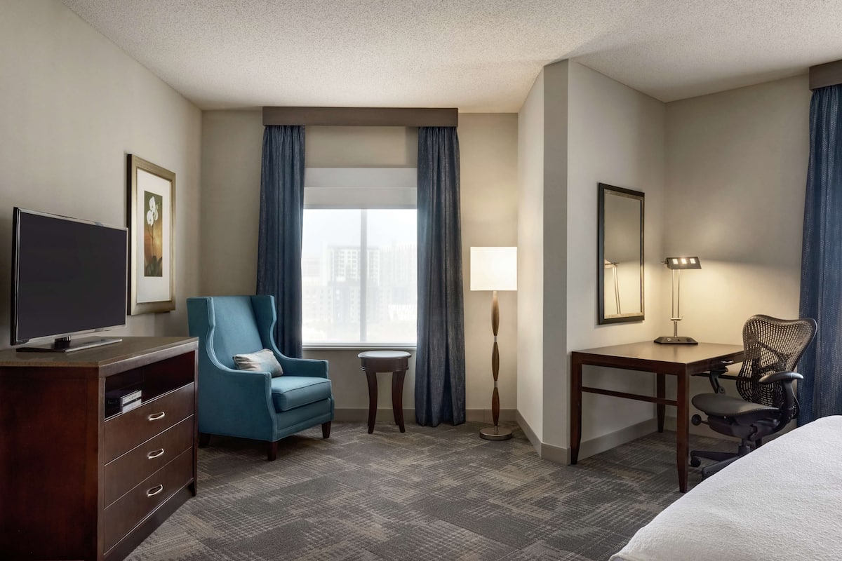 Habitación de hotel con cama, escritorio, silla, televisión, lámpara y ventana con cortinas.