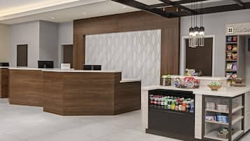 A hotel reception area with a wooden reception desk, a patterned wall, and a shelf with snacks and beverages.