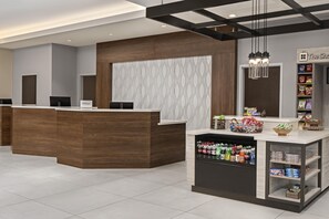Uma área de recepção de hotel com balcão de madeira, parede estampada e uma estante com lanches e bebidas.