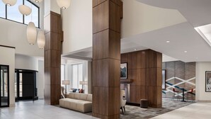 Um hall amplo com design moderno, destacando um grande sofá, uma mesa de centro e uma lareira. Possui pilares de madeira e uma janela grande que proporciona iluminação natural.