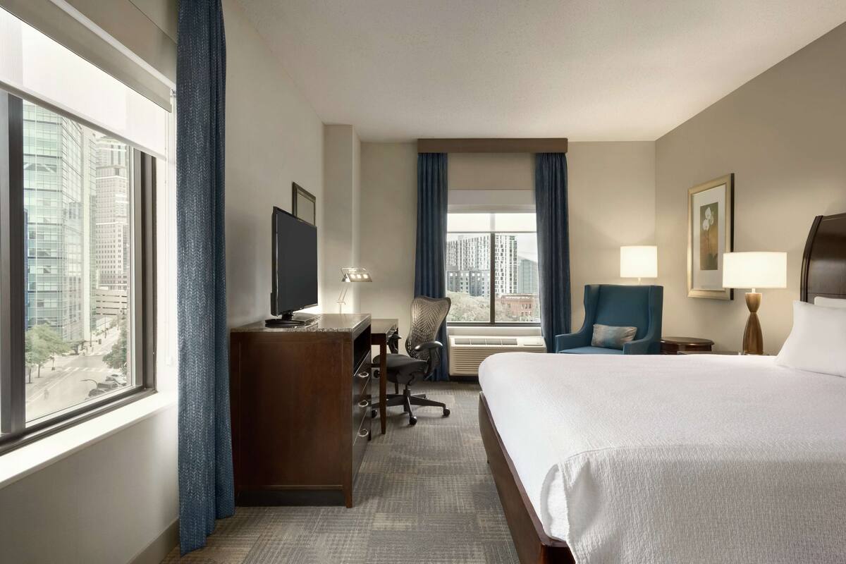 Una habitación de hotel con una cama grande, un escritorio con computadora, una silla, un televisor y una ventana con cortinas.