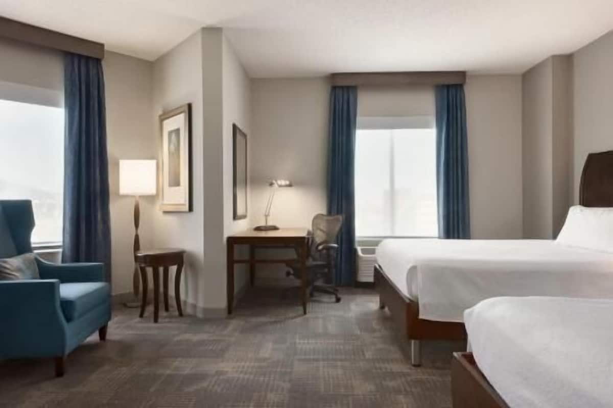 Una habitación de hotel con cama, escritorio, silla, lámpara y ventana con cortinas.