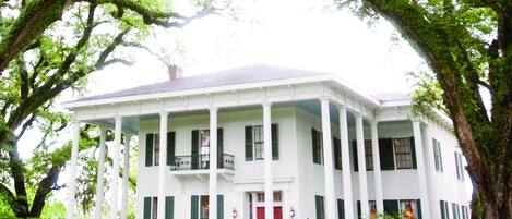 Una casa de estilo colonial blanca de dos pisos, con un porche central sostenido por columnas, rodeada de árboles verdes y frondosos.