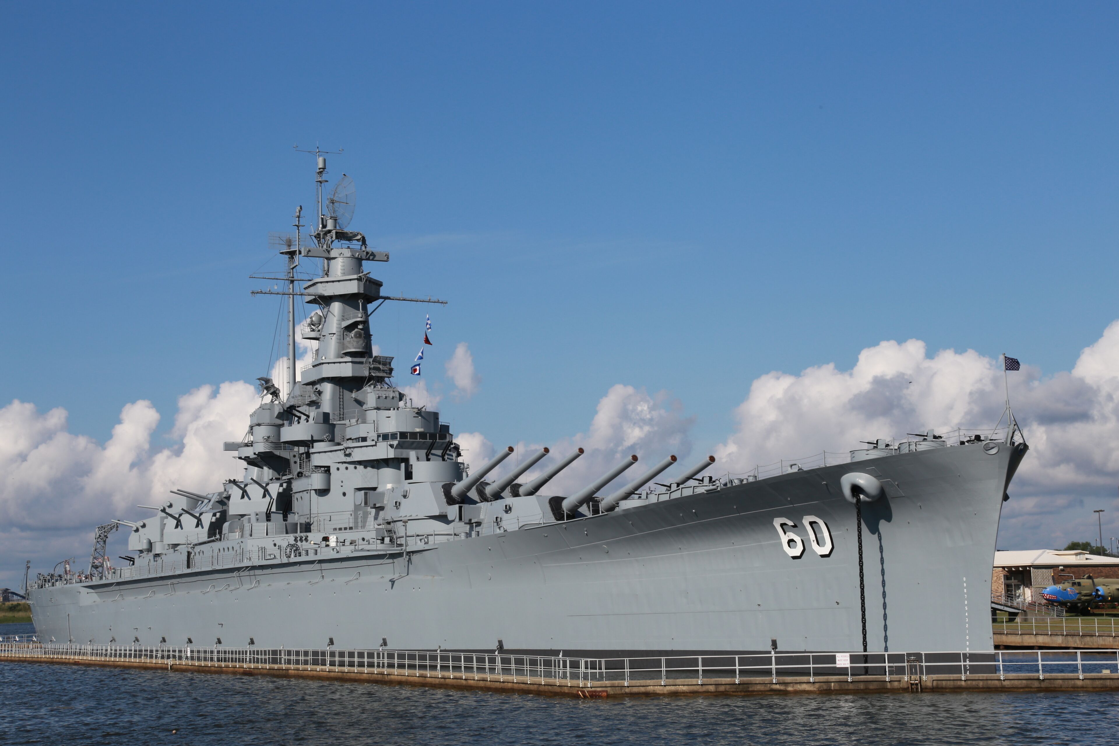 A large naval ship docked at a port with a clear sky and clouds in the background.