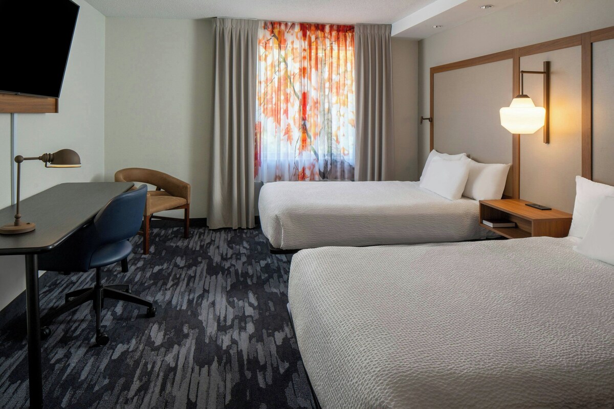 Une chambre d’hôtel avec deux lits, un bureau, une chaise, une télévision et une fenêtre avec des rideaux.