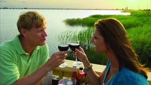 Deux personnes trinquent avec des verres de vin à une table près d’une étendue d’eau, avec une zone herbeuse et une vue lointaine sur les gratte-ciel de la ville.