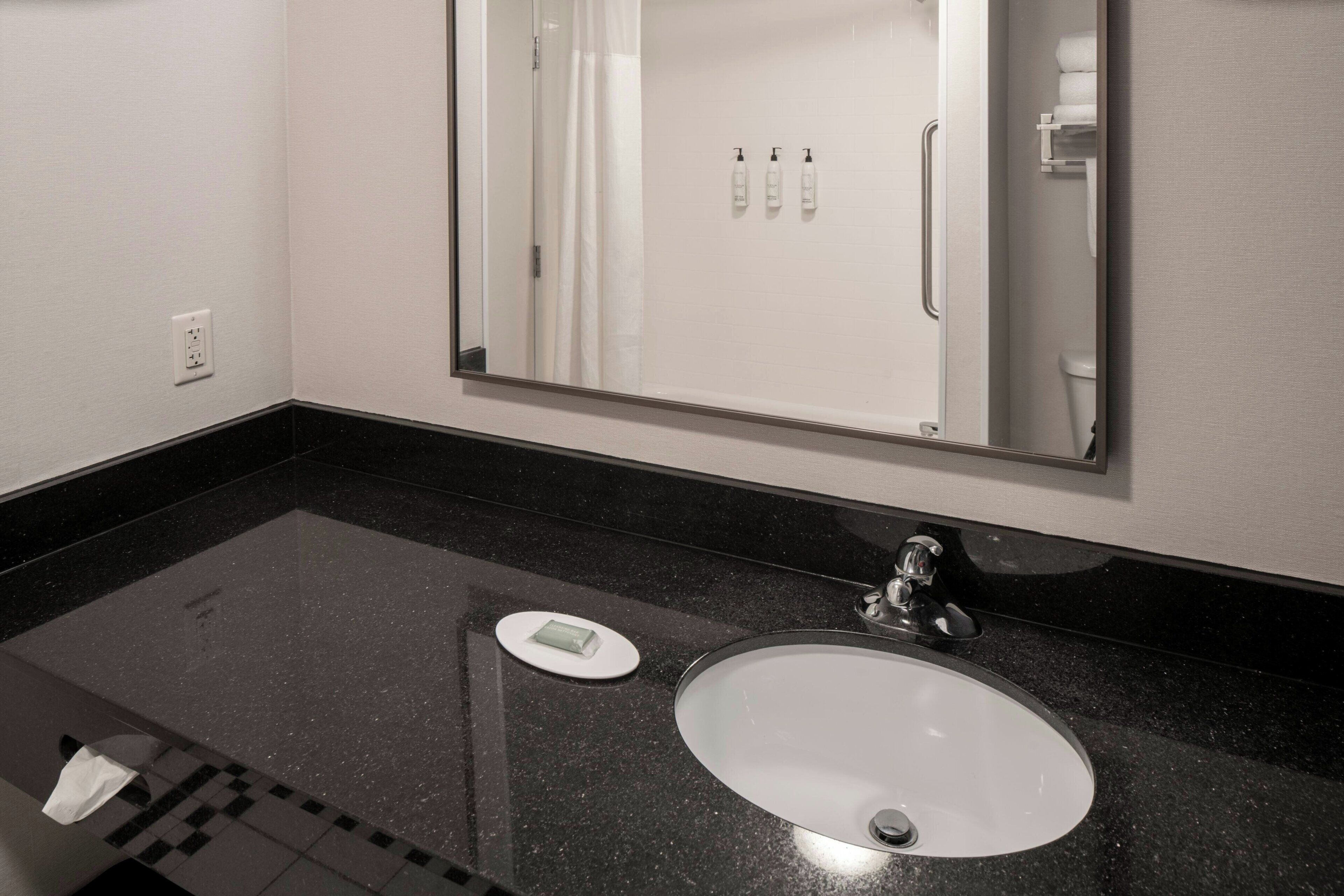 Un baño con encimera negra, lavabo blanco, espejo y dispensador de toallas.