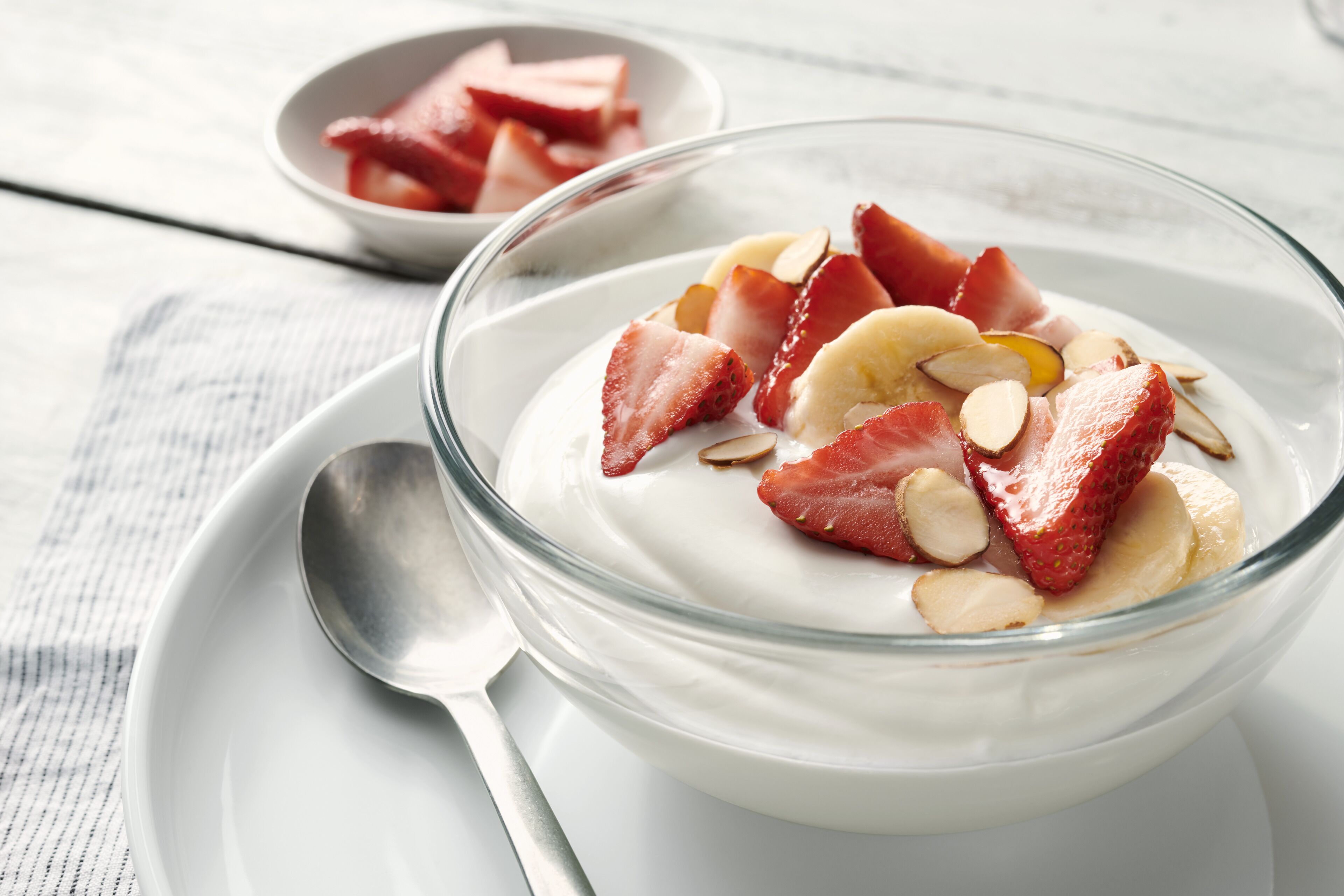 A bowl of yogurt topped with strawberries and almonds on a white plate with a spoon beside it.