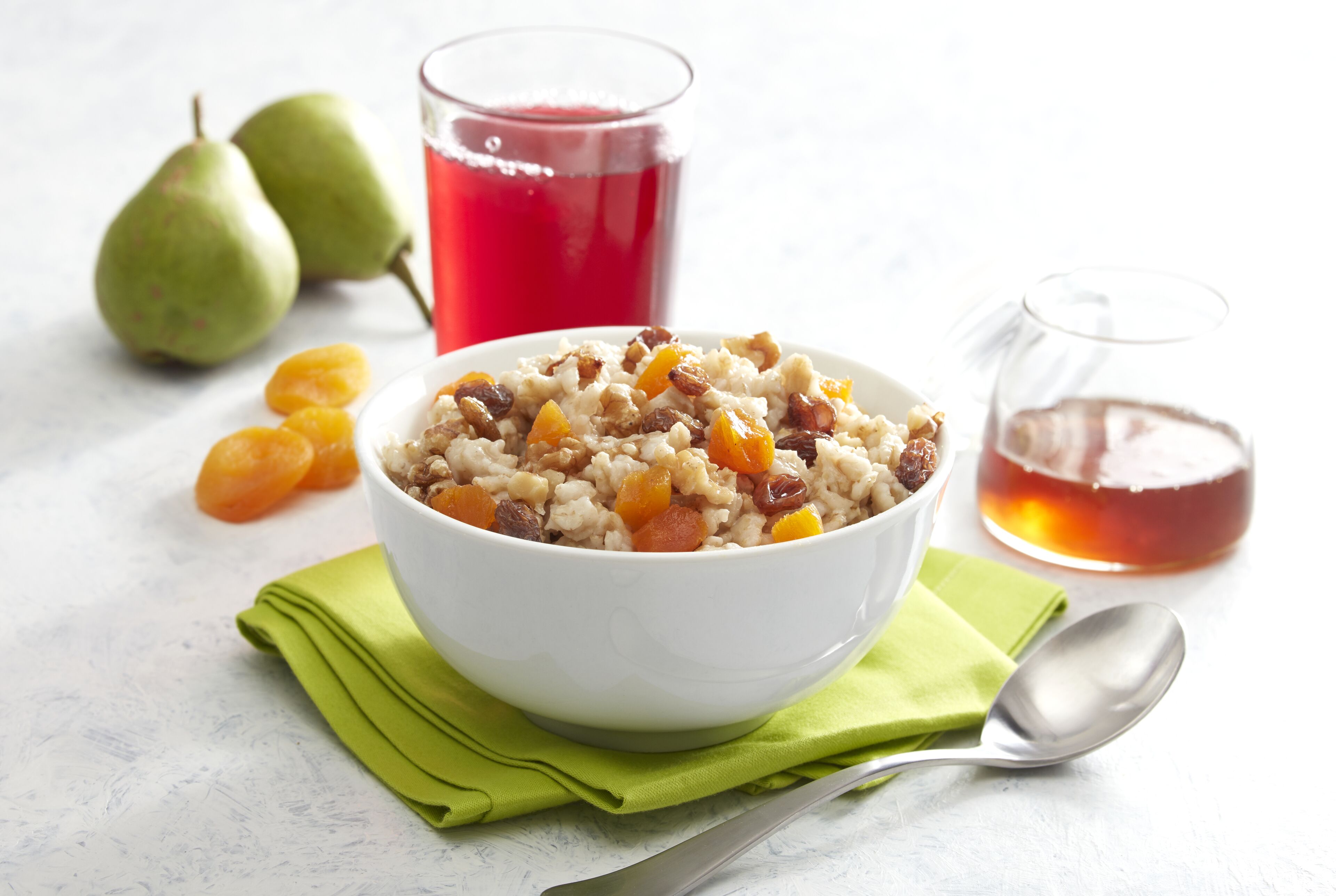 A bowl of oatmeal with raisins and apricots, a glass of red juice, and two pears on a white surface.