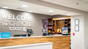 A hotel reception area with a 'Welcome to the Creole State' sign, a Hilton Honors sign, a self-service kiosk, and a snack bar. - Hampton Inn Gonzales (Gonzales)