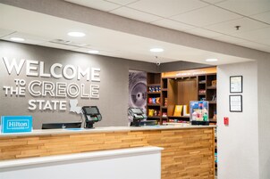 Zona de recepción de hotel con un cartel que dice 'Welcome to the Creole State', un letrero de Hilton Honors, un quiosco de autoservicio y una barra de aperitivos.