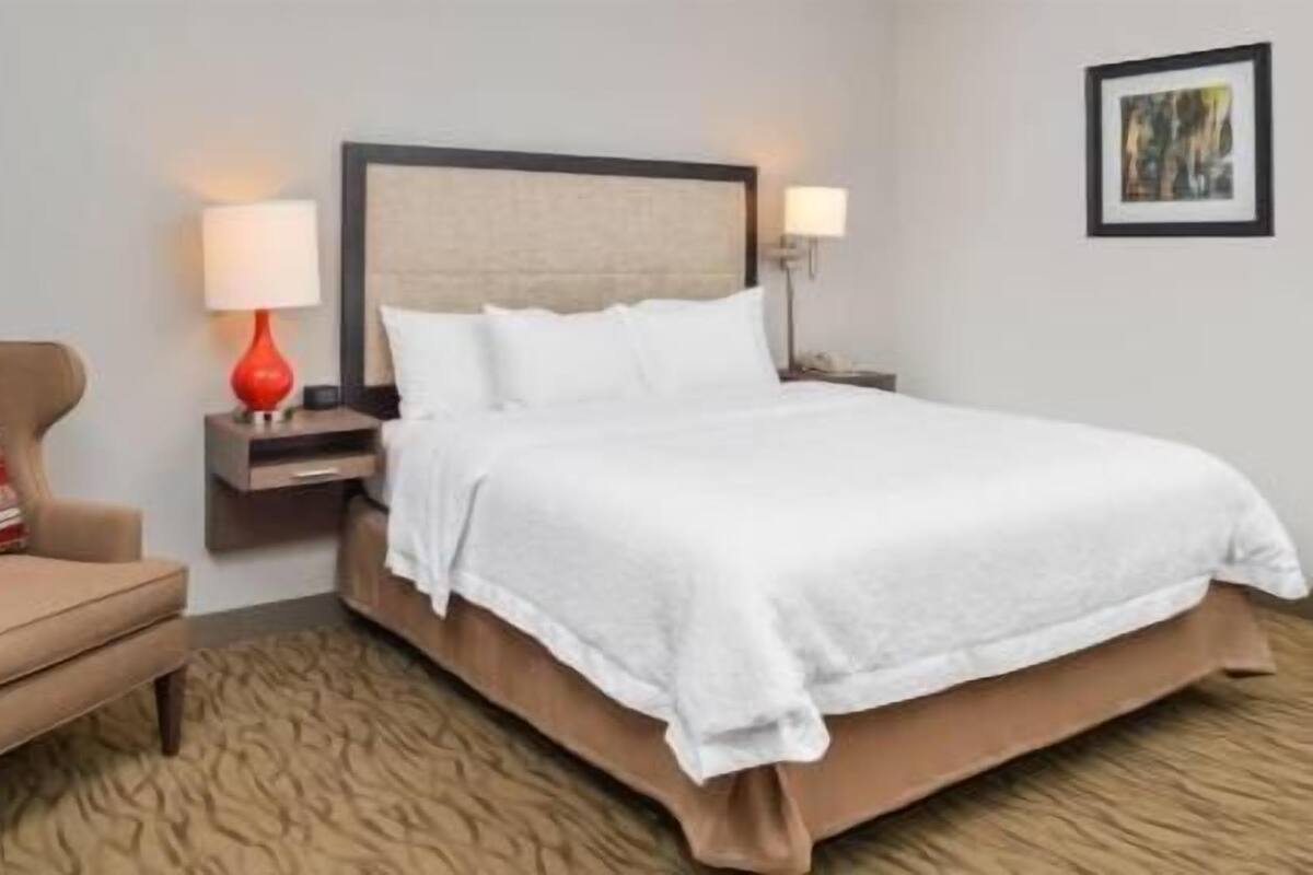 Quarto de hotel com cama, cadeira, mesa de cabeceira e abajur.