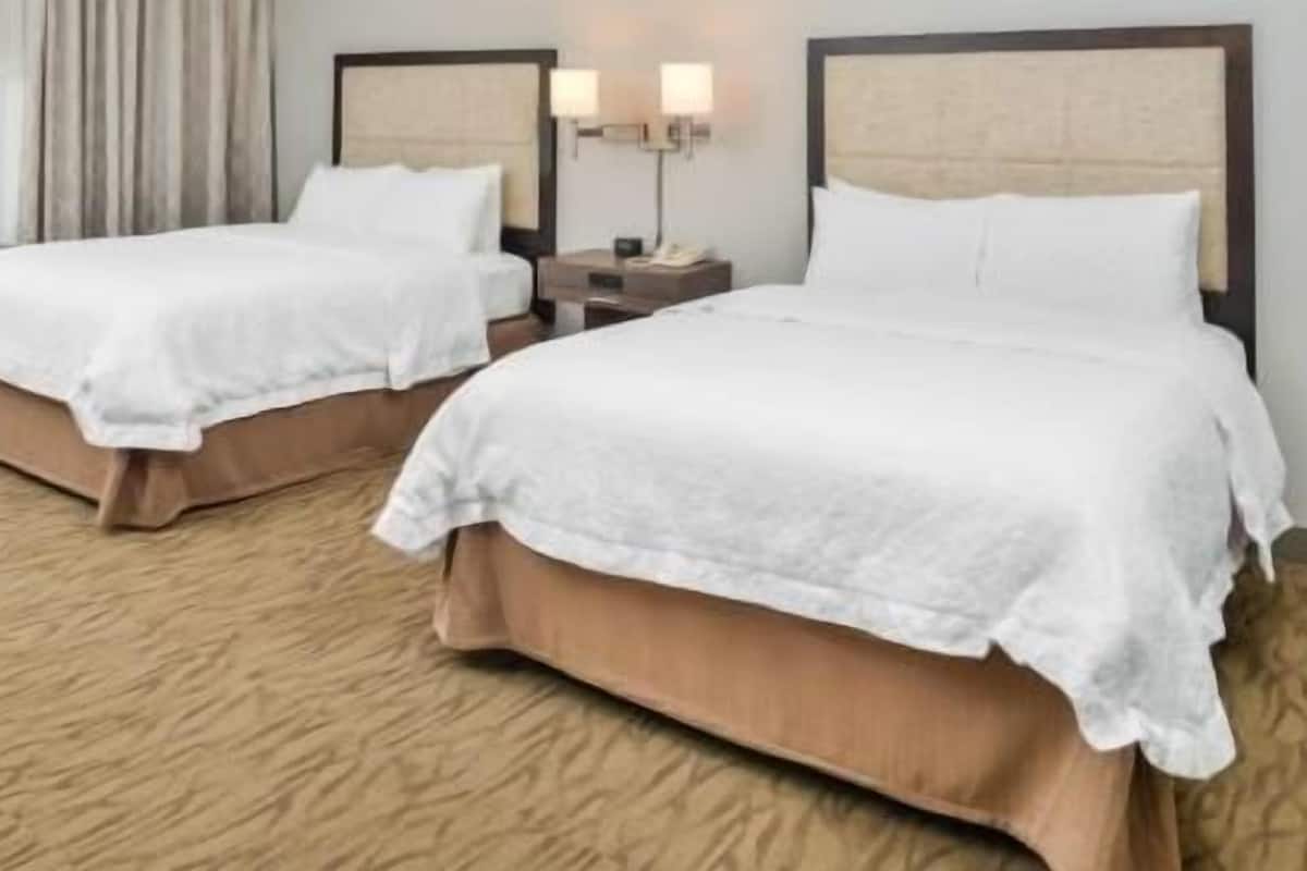 Two double beds with white linens, a wooden headboard, a nightstand with a lamp, and a carpeted floor.