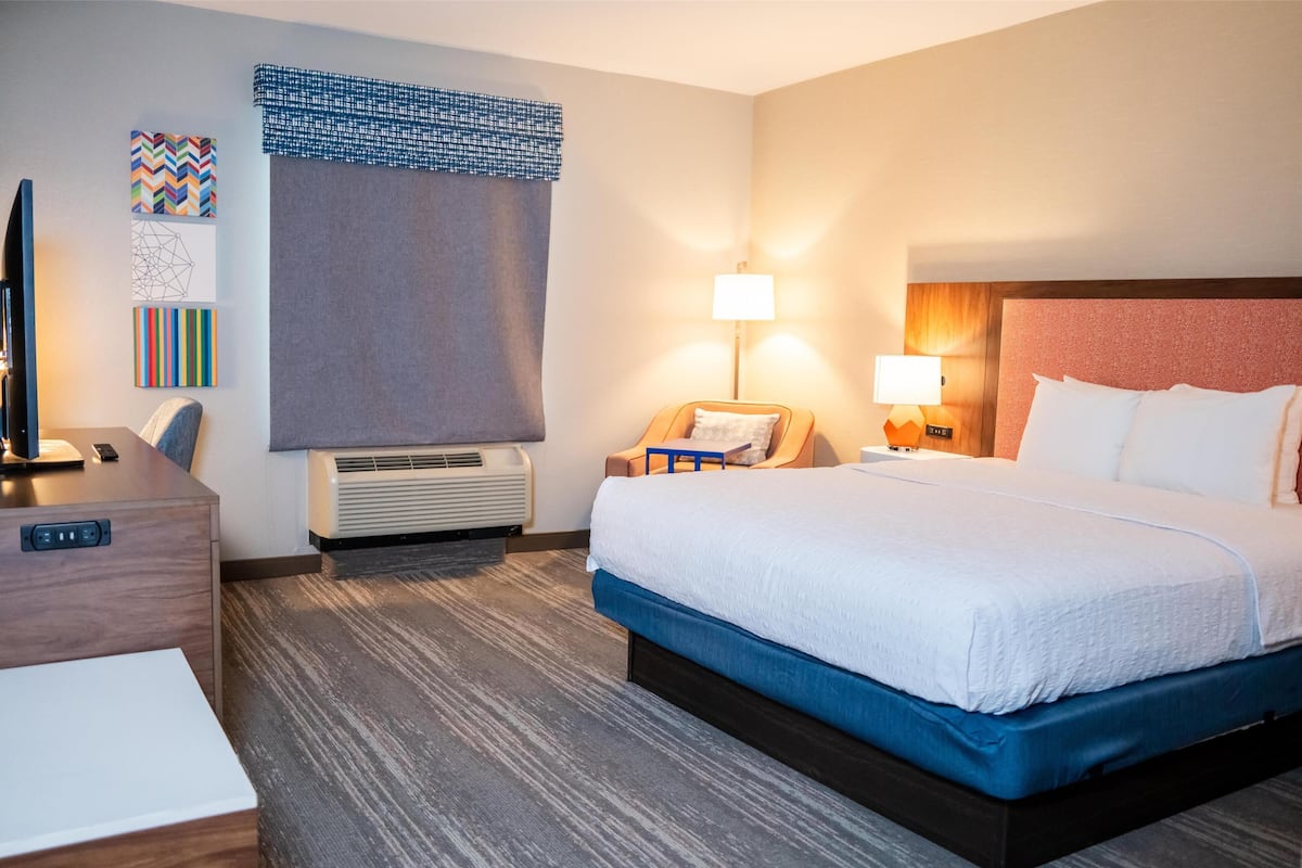 A hotel room with a bed, bedside tables, a desk, a chair, a lamp, and wall art.