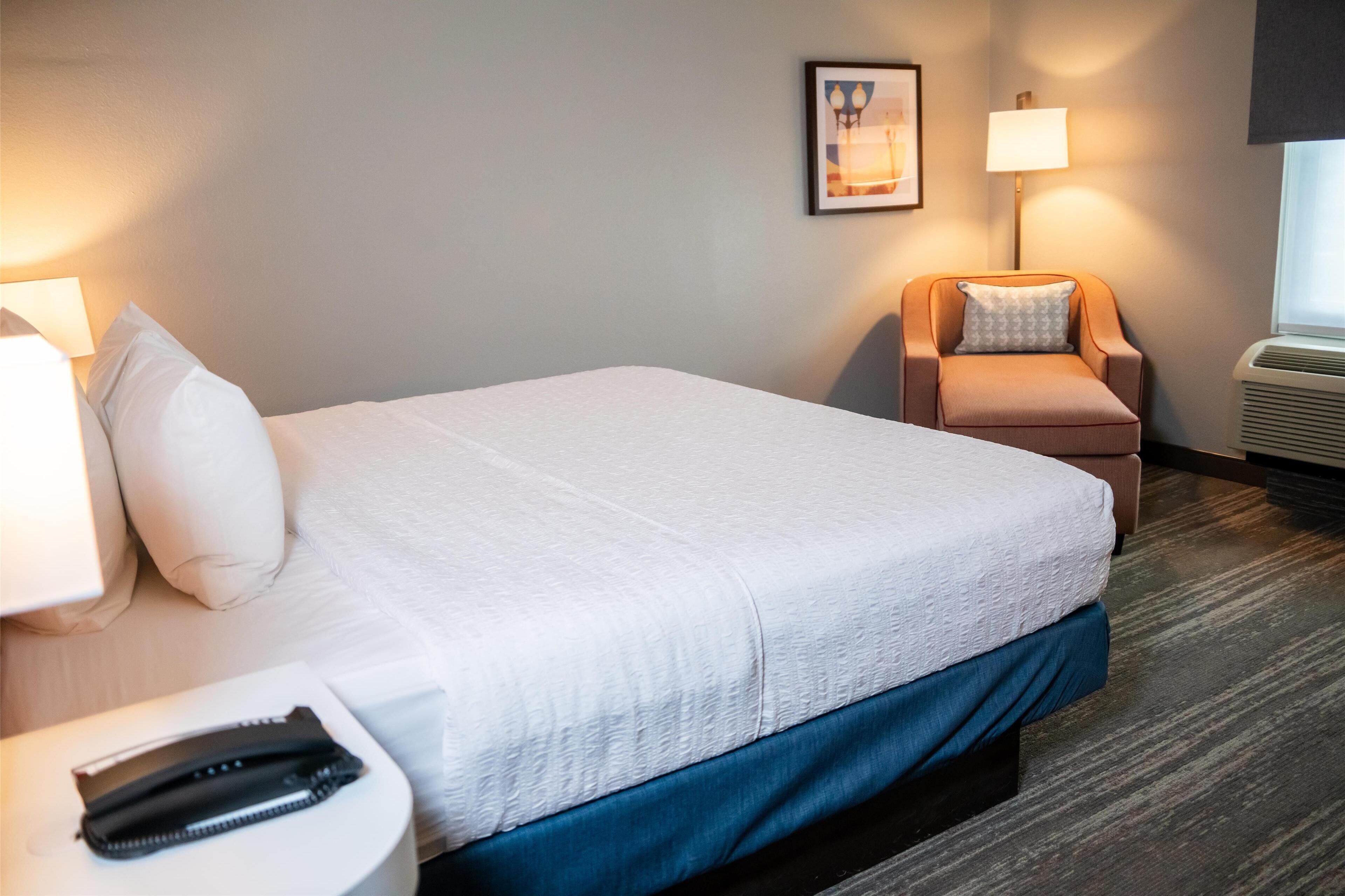 A hotel room with a bed, a chair, a lamp, a phone, and a picture on the wall.