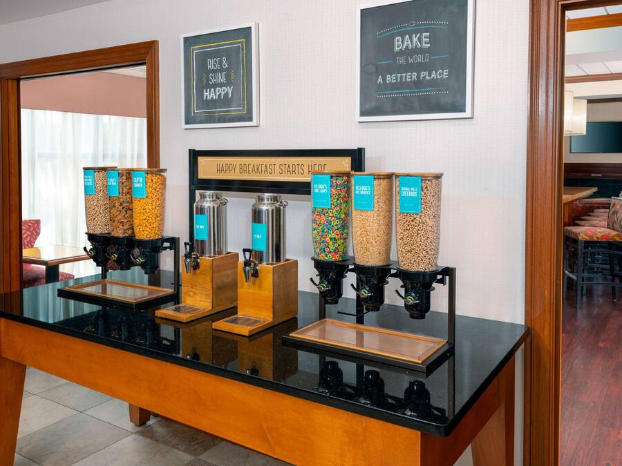 A breakfast bar with various food dispensers, a sign reading "HAPPY BREAKFAST STARTS HERE", and framed motivational quotes on the wall.